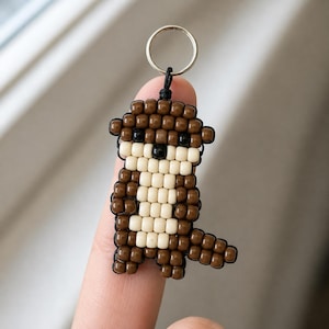 May include: A keychain crafted from brown and cream-colored plastic pony beads arranged to form the shape of an otter. The design features two small black beads for eyes and one black bead for a nose, all connected to a silver metal ring.