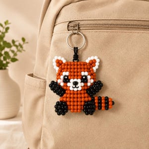 May include: A red panda keychain crafted from small, round, orange, white, and black beads. The figure features a striped tail, black limbs, and a white face with black eyes and nose. It is attached to a silver-toned metal split ring.