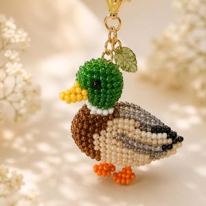 Könnte beinhalten: Ein Enten-Schlüsselanhänger aus kleinen, runden Perlen in den Farben Grün, Gelb, Braun, Creme, Grau und Orange. Die Ente hat einen grünen Kopf, einen gelben Schnabel, eine braune Brust und orangefarbene Füße und ist an einem goldenen Karabiner befestigt.