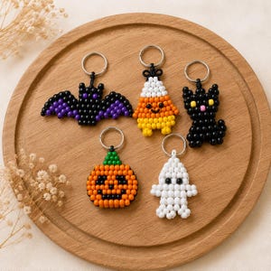 May include: Five Halloween-themed keychains made of small plastic beads. The set includes a black bat with purple wings, a candy corn figure, a black cat with yellow eyes, an orange pumpkin with a green stem, and a white ghost with black eyes.