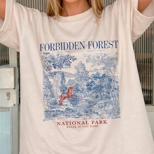Pode incluir: Camiseta creme com um gráfico azul e vermelho de uma cena de floresta e o texto "FORBIDDEN FOREST NATIONAL PARK ENTER IF YOU DARE". A camiseta tem mangas curtas e um corte folgado.