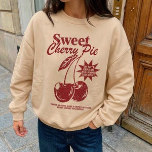 Pode incluir: Uma camisola bege de gola redonda com um gráfico vermelho escuro de duas cerejas, o texto Sweet Cherry Pie, uma estrela com o texto OH SH*T GRAHAM YOU KNOW THIS ONE e o texto TASTES SO GOOD, MAKE A GROWN MAN CRY.