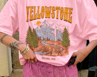 May include: A light pink short-sleeved t-shirt featuring a graphic print of Yellowstone National Park with mountains, pine trees, a river, and a bison. The text YELLOWSTONE is printed in large orange letters above the landscape illustration.