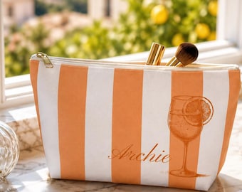 May include: A rectangular zippered pouch featuring vertical orange and white stripes. The front displays a graphic of a stemmed glass with a citrus slice and the name Archie in cursive script. The pouch contains two makeup brushes.