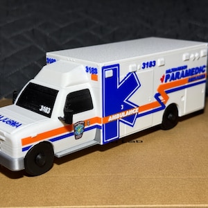 Peut inclure: Une ambulance jouet blanche avec des bandes bleues et orange, le texte PARAMEDIC et le numéro 3183. Le véhicule présente une grande lettre K bleue sur le côté, un emblème étoile de vie sur la porte et le mot AMBULANCE imprimé sur le côté.