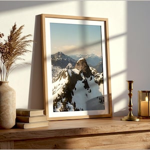May include: A framed landscape photograph titled Mountain Peaks, Vancouver, BC, depicting snow-capped mountains. The print is displayed on a wooden surface next to a textured vase with dried grass, a stack of books, a brass candle holder, and a candle.