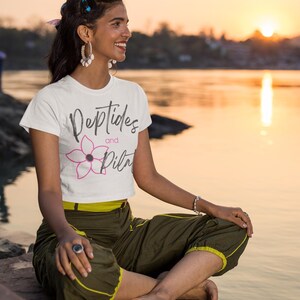 Può includere: Maglietta bianca con le parole "Peptides and Pilates" e una grafica floreale rosa. La persona indossa pantaloni verde oliva con finiture gialle ed è seduta a gambe incrociate vicino all'acqua al tramonto.