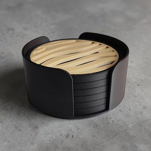 May include: A stack of circular black coasters housed in a matching black cylindrical holder. The top coaster features a light beige surface with a series of parallel, curved, wave-like cutouts that reveal the black coaster underneath.