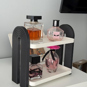 May include: A two-tiered white tray organizer with black arched side supports. The top shelf holds a square glass perfume bottle with amber liquid and a round glass bottle with pink liquid. The bottom shelf holds a square bottle and a pink-topped bottle.