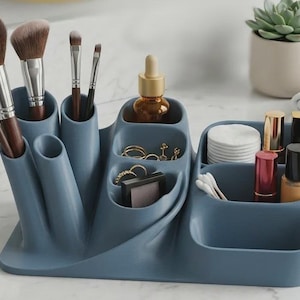 May include: A slate blue multi-compartment vanity organizer featuring various slots for makeup brushes, a small amber glass dropper bottle, gold rings, a compact, a stack of white cotton rounds, cotton swabs, and several lipstick tubes.