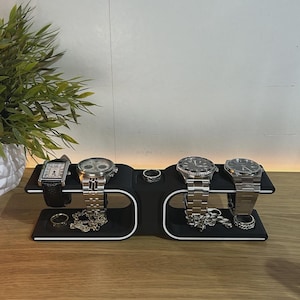 May include: A black watch and jewelry organizer stand with white trim, featuring two horizontal tiers. The stand holds four wristwatches with metal and leather bands, two silver-toned rings, and two silver-toned chain bracelets displayed on the base.