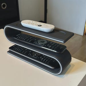 Op de afbeelding: Een donkergrijze, S-vormige afstandsbedieningshouder met twee niveaus. Het bovenste niveau bevat een witte streaming-afstandsbediening, terwijl het middelste en onderste niveau elk een zwarte televisie-afstandsbediening bevatten.