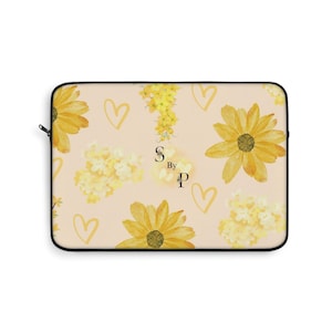 Op de afbeelding: Een laptoptas met een bloemenpatroon. Het heeft gele zonnebloemen, hartvormen en de tekst "S By P" op een licht perzikkleurige achtergrond. De tas heeft een zwarte rits en rand.