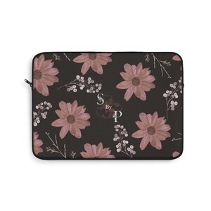 Op de afbeelding: Een zwarte laptoptas met een bloemenpatroon van grote, stoffige roze bloemen en witte bessen takjes. Het ontwerp bevat de tekst "S By P". De tas heeft een zwarte ritssluiting.
