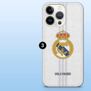 Op de afbeelding: Een witte smartphonehoes met het embleem van voetbalclub Real Madrid in geel, blauw en rood, met verticale lichtgrijze strepen en de tekst &iexcl;HALA MADRID! in het zwart onderaan gedrukt. De hoes heeft een uitsparing voor een drievoudige camera.