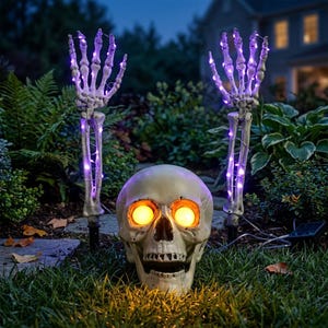 May include: A Halloween yard decoration featuring a realistic off-white human skull with glowing yellow eyes positioned between two skeletal arms. Each arm is upright, featuring articulated fingers wrapped in purple LED string lights.