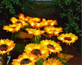 May include: A collection of glowing artificial sunflowers, featuring vibrant yellow petals and dark brown centres. Each flower is adorned with tiny, warm white lights. The sunflowers are set against a backdrop of green leaves, creating a garden setting.