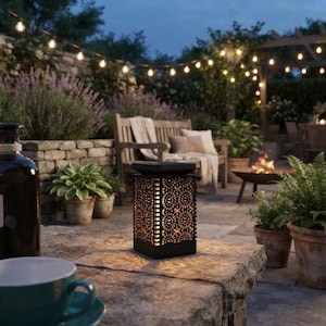 May include: A square black metal solar lantern with a decorative laser-cut floral pattern on all four sides, emitting a warm internal glow. The lantern sits on a stone surface next to a teal ceramic mug, with a garden patio setting in the background.