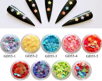 May include: Black nail tips adorned with pastel flower-shaped glitter. Below, small clear jars filled with various colors of flower-shaped glitter, including iridescent, yellow, blue, pink, and red. Each jar is labeled with a "GD55-" followed by a number.