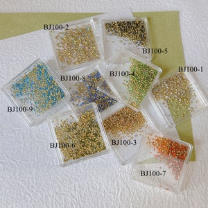 Könnte beinhalten: Sortiment kleiner, farbiger Perlen in transparenten quadratischen Behältern. Die Perlen sind in verschiedenen Farben erhältlich, darunter Gold, Grün, Blau und Orange. Jeder Behälter ist mit "BJ100-" und einer Nummer beschriftet.