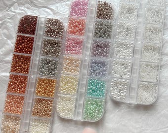 Pode incluir: Três recipientes de plástico transparente cheios de pequenas contas redondas de várias cores. As contas são em tons de castanho, rosa, branco e verde. Uma única unha com um desenho de contas está no canto inferior direito.