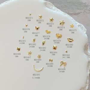 Op de afbeelding: Assortiment van gouden nagelcharms in verschillende vormen en maten, waaronder sterren, harten, bladeren, strikken en geometrische vormen. Elke charme is gelabeld met een productcode en een maat in millimeters.