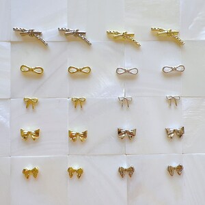 May include: A set of 18 gold and silver bow-shaped nail charms arranged in a grid pattern on a white background.