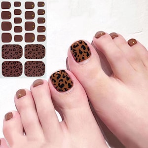 Könnte beinhalten: Braune und schwarze Leoparden-Nagelaufkleber für Zehennägel. Die Aufkleber werden auf die Zehennägel der Füße einer Person geklebt.