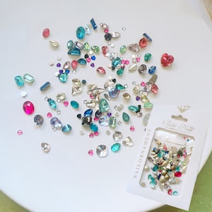Op de afbeelding: Een verzameling kleurrijke nail art strass steentjes in diverse vormen, waaronder harten, vierkanten en ovalen. De edelstenen zijn verspreid op een witte ondergrond, met een verpakking Best Nail nail ornaments.