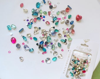 May include: A collection of colorful nail art rhinestones in various shapes and sizes, including hearts, squares, and ovals. The gems are scattered on a white surface, with a package of Best Nail nail ornaments in the corner.