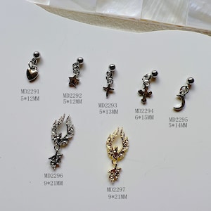 Op de afbeelding: Assortiment van 10 zilveren en gouden oorhangers met verschillende bedels, waaronder een hart, een ster, een kruis, een halve maan en vleugels. Elke oorhanger is gelabeld met een productcode en afmetingen in millimeters.