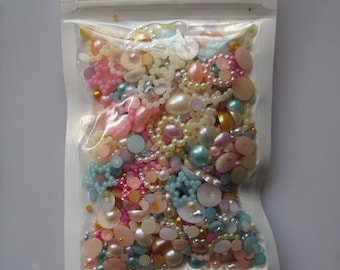 Pode incluir: Um saco de plástico transparente cheio de uma variedade de contas, pérolas e botões em cores pastel. As contas são em tons de rosa, azul, branco e dourado.