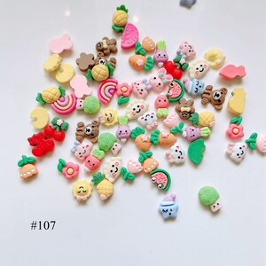 May include: Assortment of colorful resin flatback embellishments featuring various cute cartoon characters and fruit designs.  The designs include a rainbow, watermelon, pineapple, strawberries, carrots, and a variety of animals. #107