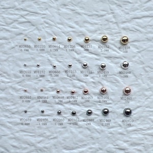 May include: A collection of small, round beads in various metallic finishes, including gold, silver, and rose gold. Sizes range from 0.8mm to 3mm, with product codes printed below each bead. The beads are arranged in rows on a light-colored surface.