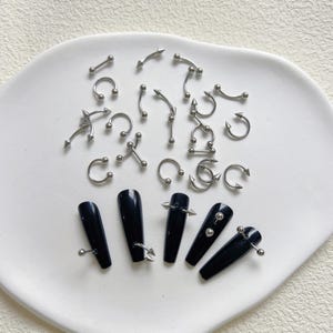 May include: An assortment of silver body jewellery, including curved barbells, circular barbells, and other piercing styles. Also, black acrylic nails with silver studs and ball accents are displayed on a white plate.