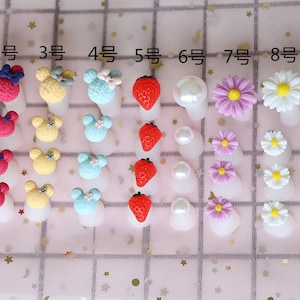 May include: Various nail art decorations, including cartoon mouse heads, strawberries, pearls, and flowers, displayed on white nail tips. Numbers 1 to 9 are above each design. The background is a pink fabric.