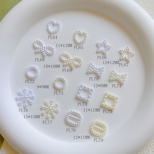 Op de afbeelding: Assortiment van witte parelversieringen in verschillende vormen en maten, waaronder harten, sterren, cirkels, oneindigheidssymbolen en strikken. Elke versiering heeft een bijbehorende productcode en een maat in millimeters.