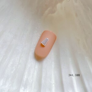 Puede incluir: Un pequeño charm para uñas rectangular, blanco e iridiscente, con una parte superior ligeramente puntiaguda. El charm mide 3 por 4,5 milímetros.