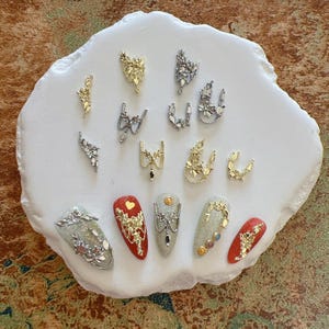 Op de afbeelding: Een assortiment nail art decoraties en versierde nagels. De decoraties omvatten gouden en zilveren bloemmotieven, strikken en andere versieringen. Verschillende nagels zijn rood, zilver en grijs geschilderd, met gouden en zilveren accenten.
