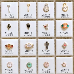 Puede incluir: Surtido de 12 pequeños abalorios decorativos para el arte de las uñas. Los abalorios están hechos de metal y presentan varios diseños, incluyendo llaves, relojes, cruces, estrellas y flores. Los abalorios son de varios colores, incluyendo oro, plata, negro, blanco, rojo, azul y verde. Los abalorios están etiquetados con su tamaño y código de producto.