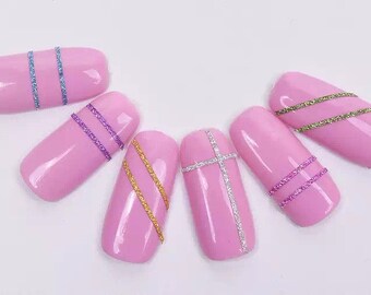 Puede incluir: Uñas artificiales rosas con detalles brillantes. Los diseños incluyen rayas horizontales en azul, morado y dorado, y una cruz plateada. Las uñas están dispuestas sobre una superficie blanca.