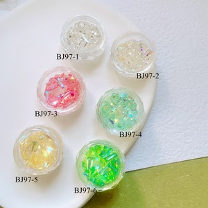 Peut inclure: Six petits pots en plastique transparent remplis de décorations d'art d'ongle colorées et iridescentes. Les pots sont étiquetés BJ97-1 à BJ97-6 et contiennent diverses couleurs, dont le transparent, le rose, le vert et le jaune. Les pots sont sur une surface blanche.