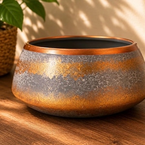 May include: A wide, low-profile bowl with a dark grey, textured exterior featuring horizontal bands of metallic gold leaf finish. The rim is accented with a smooth, polished copper-coloured band, and the interior of the bowl is a solid dark grey.