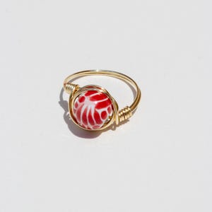 May include: A gold-toned wire-wrapped ring featuring a single round bead. The bead is red with a white marbled or floral-like pattern. The metal wire is coiled around the base of the bead to secure it to the thin, circular gold-toned band.