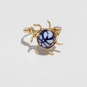 May include: A ring featuring a central round bead with a dark blue and white floral pattern, encased in a gold-toned wire setting. The wire is wrapped around the bead and extends into several curved, decorative prongs radiating outward.