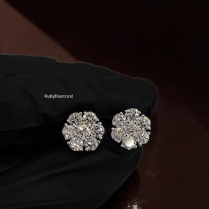 May include: A pair of stud earrings featuring a cluster of seven round brilliant-cut clear stones arranged in a floral pattern. The stones are set in a metallic base, and the text RubyDiamond is visible near the earrings on the dark background.