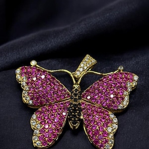 May include: A gold butterfly brooch, adorned with pink and white gemstones. The wings are set with pink stones, edged with white stones. The body is black, and the brooch has a pin fastening. The text "RubyDiamond" is at the bottom.