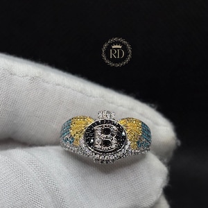 May include: A silver-toned ring featuring a central circular emblem with the letter B, surrounded by clear stones. The ring design includes wing-like accents decorated with yellow and blue stones, with additional clear stones along the band.