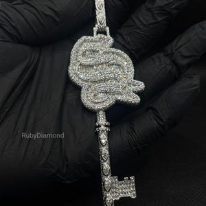 Puede incluir: Un colgante plateado con forma de llave, con una cabeza grande, ornamentada y redondeada, y un eje largo, totalmente incrustado con numerosas piedras pequeñas, claras y brillantes. El texto RubyDiamond es visible en el lado izquierdo.