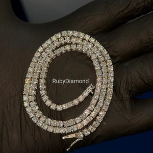 May include: A silver-toned tennis necklace featuring a continuous row of round, clear, faceted stones set in a metallic frame. The necklace is coiled in a circular shape and includes a secure box clasp closure. The text RubyDiamond is visible.